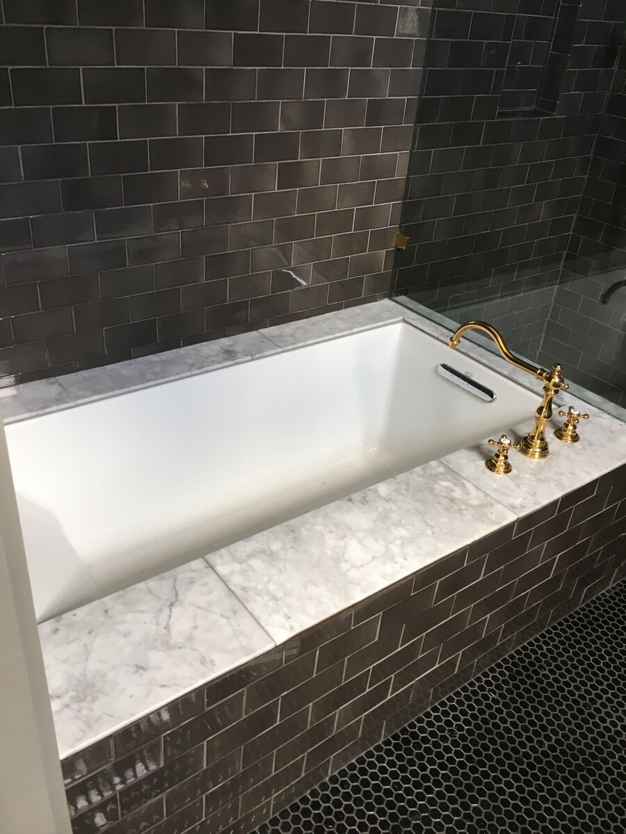 Soaking tub with black subway tile surround, Carrara marble deck, black penny tile floor, and polished brass fixtures