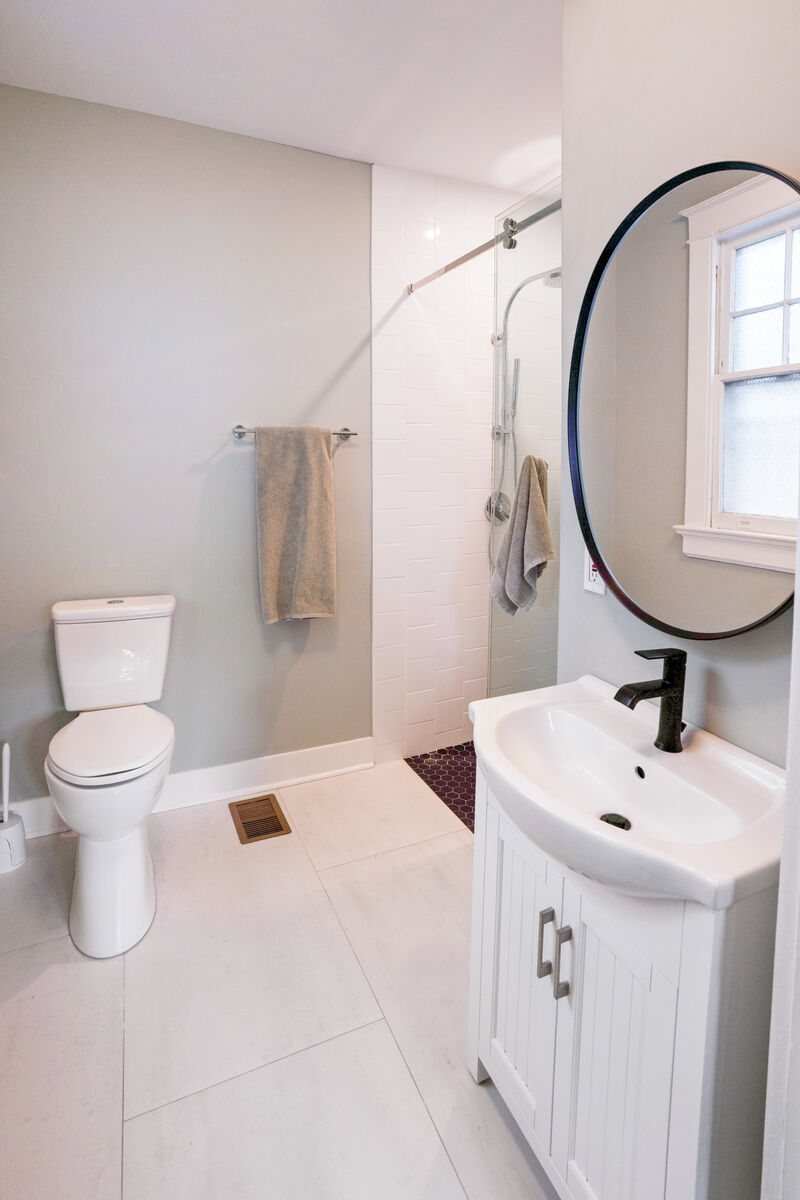 Modern white bathroom with round black-framed mirror, sliding glass shower door, hex penny tile floor, and matte black fixtures